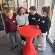 Vier Personen stehen um einen hohen roten Tisch und unterhalten sich.
