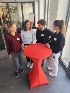 Vier Personen stehen um einen hohen roten Tisch und unterhalten sich.