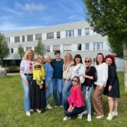 Eine Gruppe von zehn Frauen steht und kniet auf einer Wiese vor einem Gebäude und trägt bunte, traditionell bestickte Kleidung.