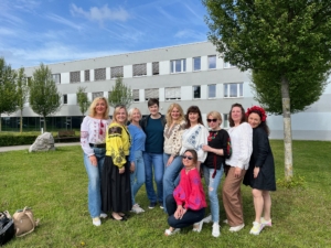 7 Eine Gruppe von zehn Frauen steht und kniet auf einer Wiese vor einem Gebäude und trägt bunte, traditionell bestickte Kleidung.