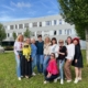 Eine Gruppe von zehn Frauen steht und kniet auf einer Wiese vor einem Gebäude und trägt bunte, traditionell bestickte Kleidung.
