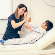 Eine Frau mit Pflegekleidung sitzt neben einem Kind auf einer medizinischen Liege und hebt die Hand zum Abklatschen.