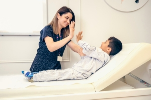 Eine Frau mit Pflegekleidung sitzt neben einem Kind auf einer medizinischen Liege und hebt die Hand zum Abklatschen.