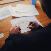 Eine Person zeichnet mit einem Stift auf einem technischen Plan, der auf einem Holztisch liegt, umgeben von weiteren Plänen und einem Stiftehalter.