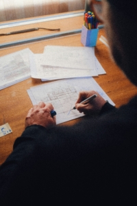 Eine Person zeichnet mit einem Stift auf einem technischen Plan, der auf einem Holztisch liegt, umgeben von weiteren Plänen und einem Stiftehalter.