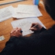 Eine Person zeichnet mit einem Stift auf einem technischen Plan, der auf einem Holztisch liegt, umgeben von weiteren Plänen und einem Stiftehalter.