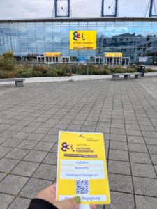 signal-2025-10-28-121806 J. Kemnitz hält ihr Namensschild für den Fürsorgetag. Im Hintergrund ist der Eingang der Messe zu sehen